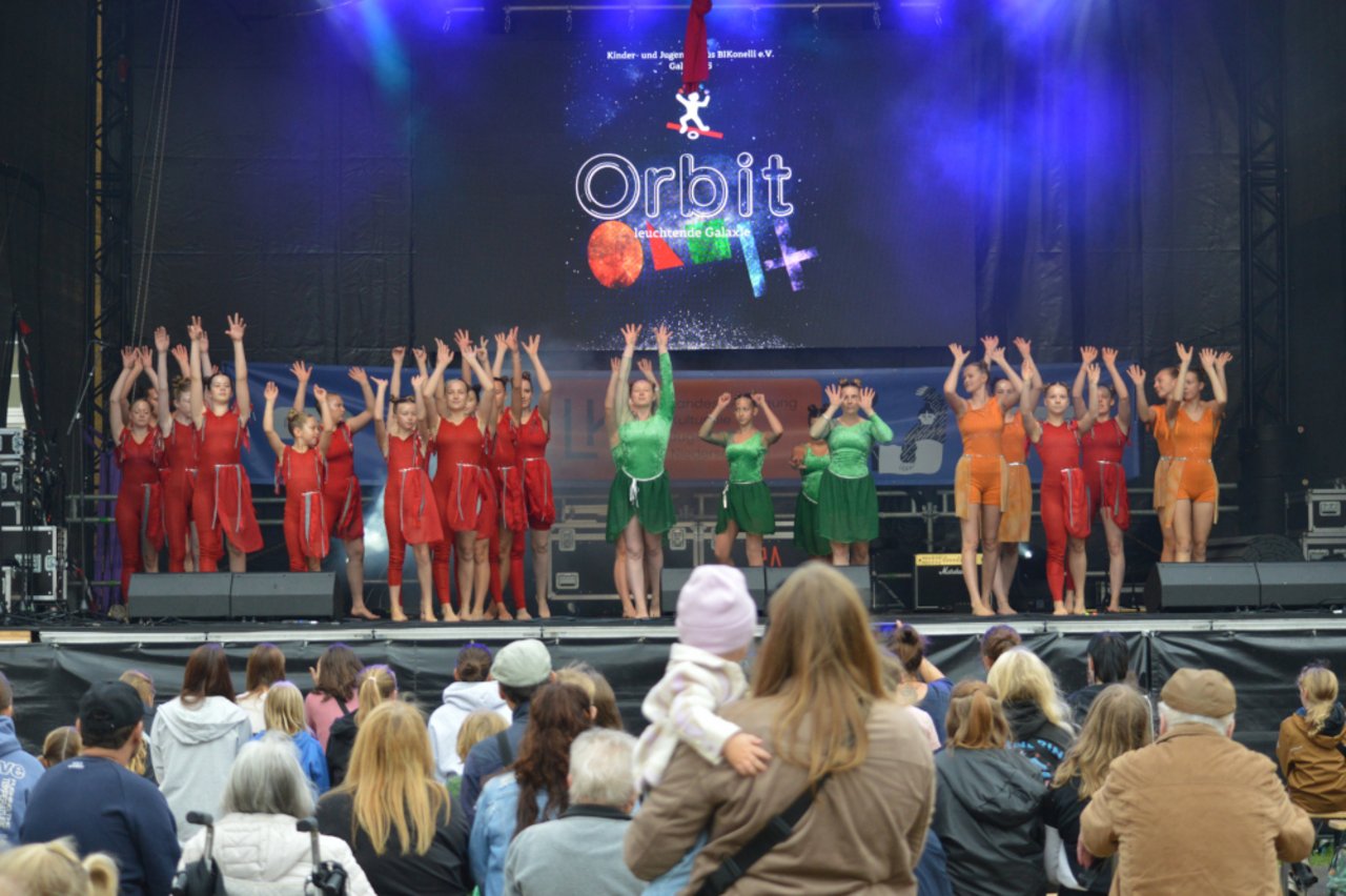 Das Bild zeigt die Kinder- und Jugendzirkusgruppe von Bikonelli e. V. aus Bad M&uuml;nder am Deister. Sie stehen mit in die H&ouml;he gereckten Armen und roten, gr&uuml;nen sowie orange farbenden Trikots auf der B&uuml;hne der LKJ vor einem gro&szlig;en Publikum.