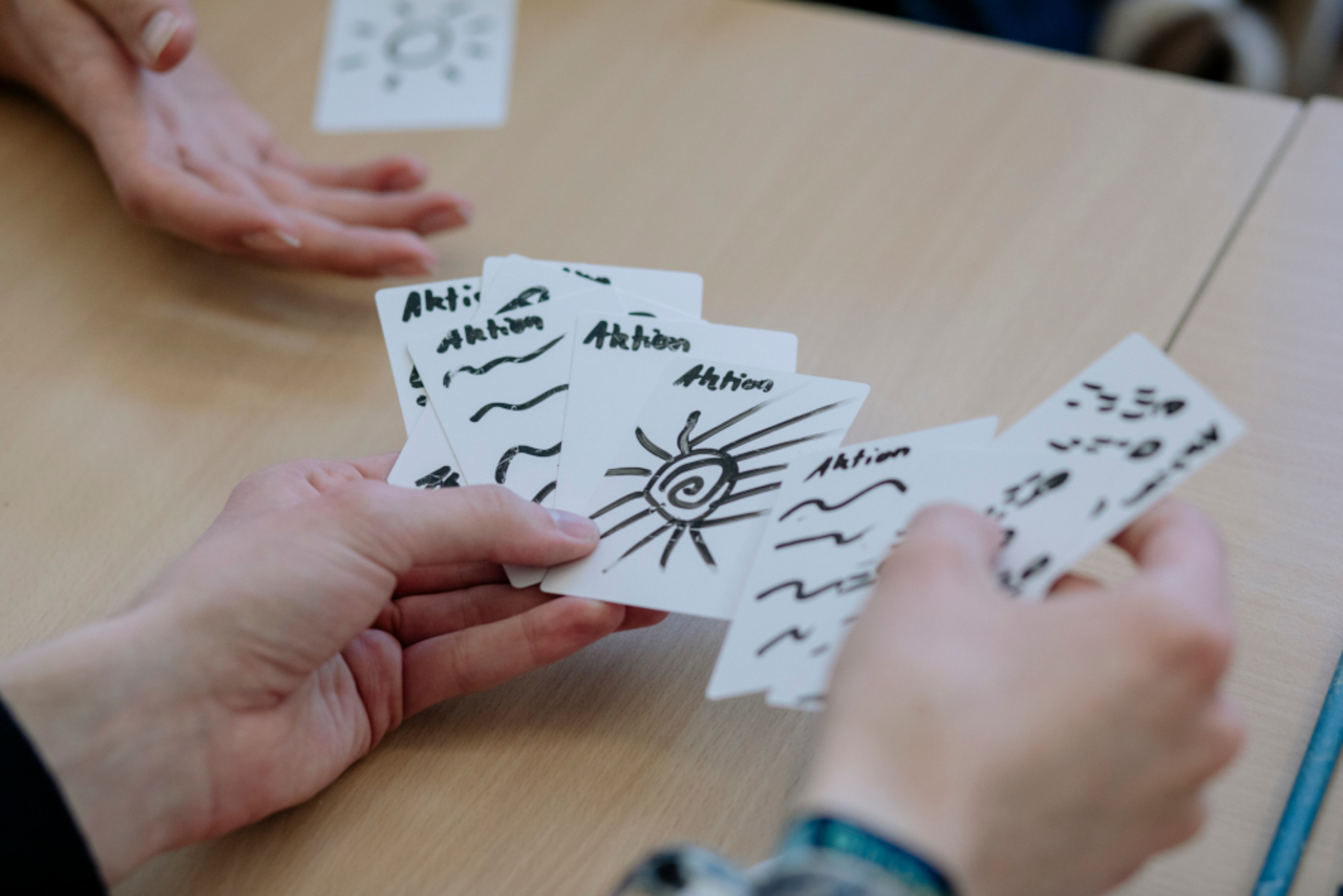Einblick in die Seminararbeit: Zu sehen ist ein Paar H&auml;nde, das wei&szlig;e Karten wie bei einem Kartenspiel ausbreitet. Auf jeder Karte ist eine Skizze zu sehen - zum Beispiel eine Sonne - und der Titel Aktion. Im oberen linken Bildrand ist eine ausgestreckte Hand zu sehen, um eine der Karten anzunehmen.