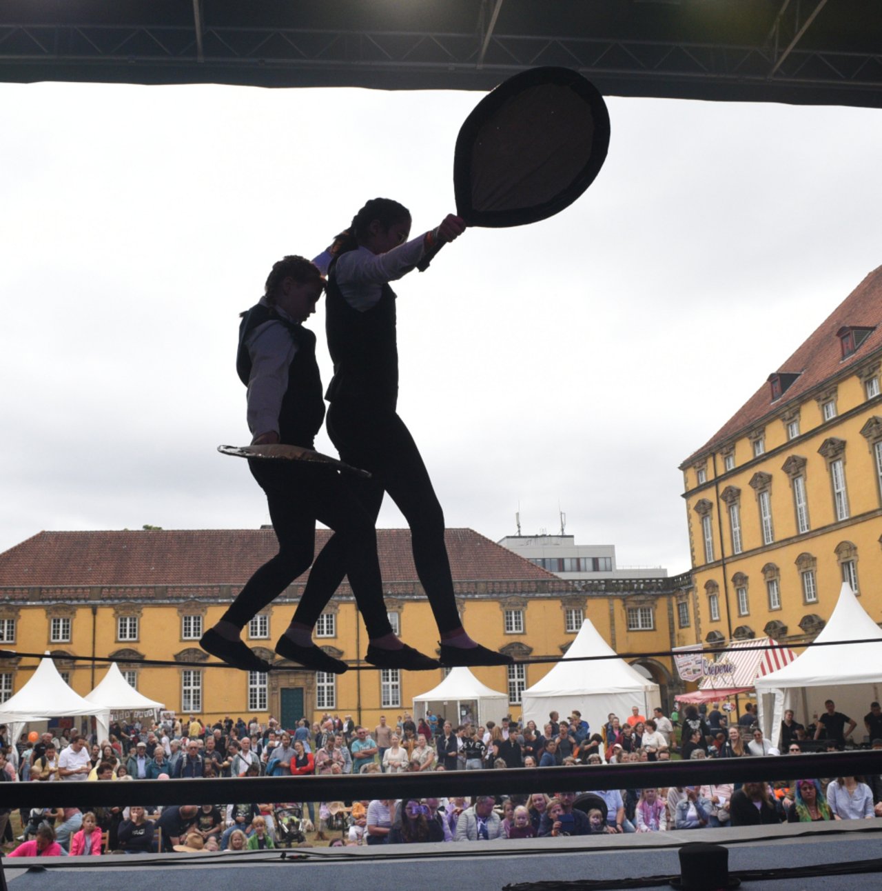 Das Foto zeigt zwei junge Seilt&auml;nzer*innen von Zirkus Knallt&uuml;te aus Celle. Sie balancieren auf der LKJ-B&uuml;hne auf einem gespannten Seil. In den H&auml;nden halten sie vermutlich Schilder. Dies ist allerdings nur schemenhaft zu erkennen, da die beiden gegen das Licht fotografiert wurden. So ist auch der Innenhof des Osnabr&uuml;cker Schlosses und das gro&szlig;e Publikum sichtbar. Die beiden Artist*innen erscheinen allerdings wie in Scherenbild-Optik und sind nur im Umriss zu erkennen. 