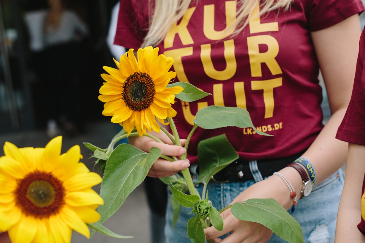 Eindr&uuml;cke der Auftaktveranstaltung: Die Jahrg&auml;nge der Freiwilligen im FSJ Kultur erhalten ein farbiges T-Shirt mit dem Aufdruck "Kulturgut". Im Jahr 2019 war es bordeauxrot mit gelbem Schriftzug. Farblich passend dazu h&auml;lt die Freiwillige eine bl&uuml;hende Sonnenblume in der Hand. 