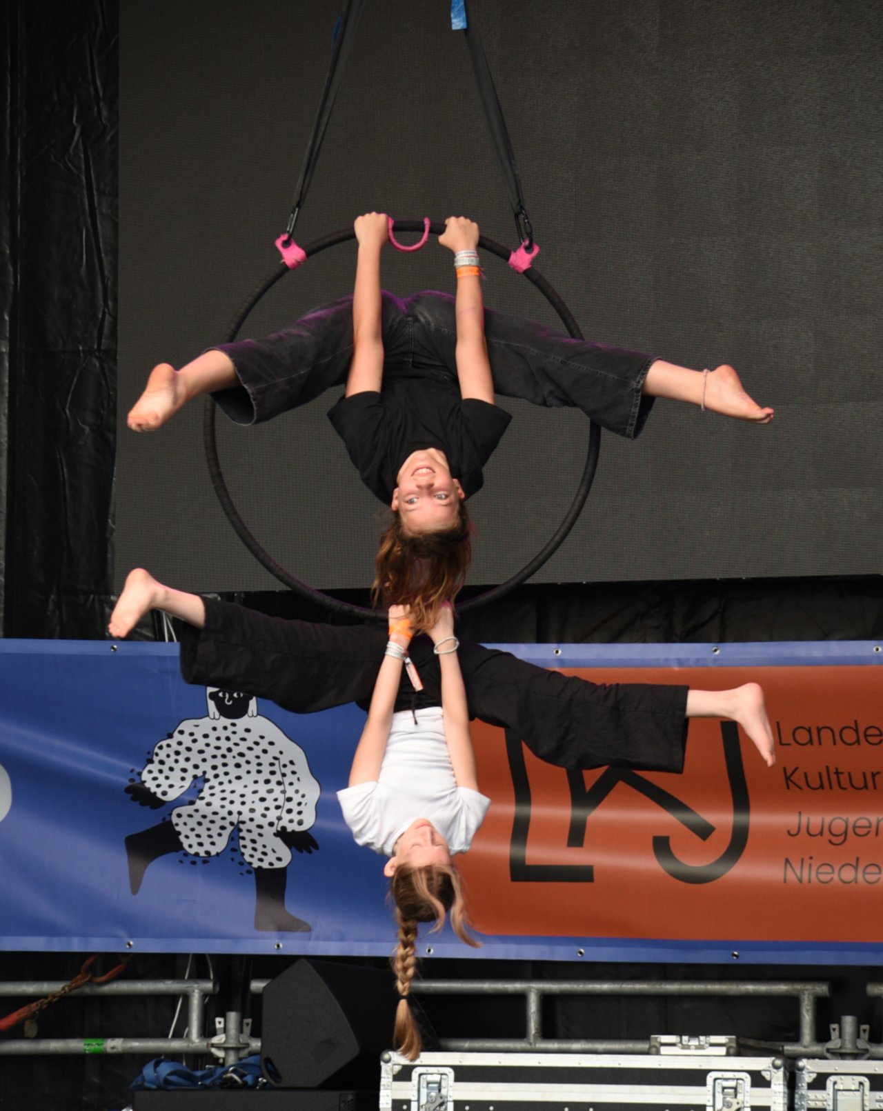 Das Bild zeigt zwei junge Artistinnen der Gruppe CirCo Hannover auf der LKJ-B&uuml;hne. Sie halten sich an einem gro&szlig;en Ring, der von der Decke h&auml;ngt, fest - eine Jugendliche am oberen Rand, die andere am unteren. Dabei h&auml;ngen sie &uuml;ber Kopf mit ausgestreckten Beinen in der Luft und l&auml;cheln.