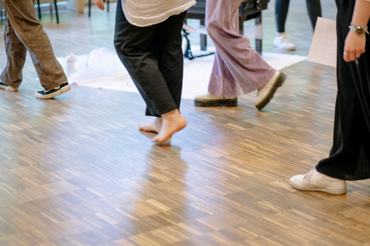 Einblick in ein Seminar: Abgebildet sind lediglich die Beine und F&uuml;&szlig;e von Menschen, die mit oder ohne Schuhe durch einen Saal schreiten. 